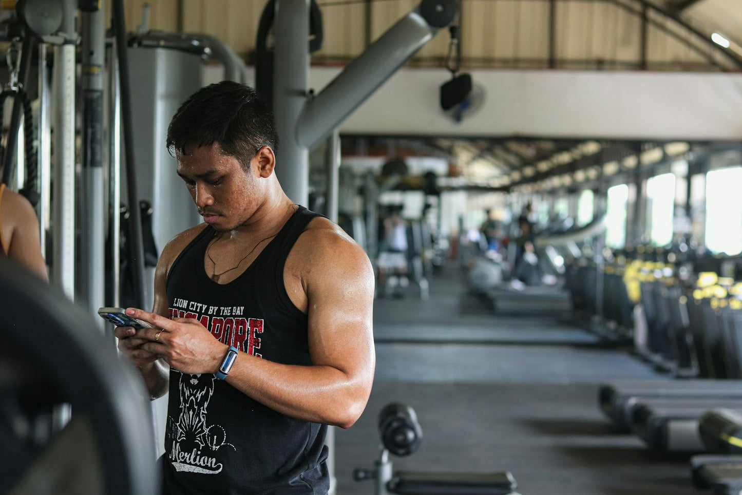 Lifter mid-workout in a gym checking his coaching app on his phone, fitness tracker on wrist
