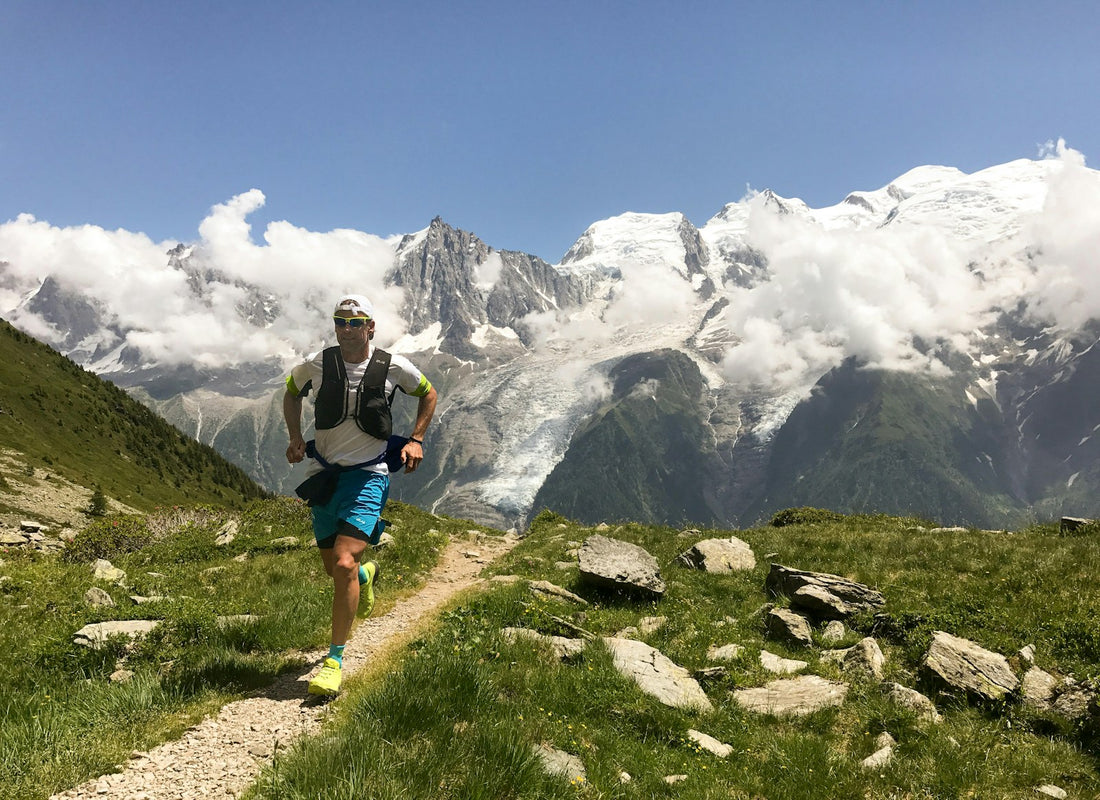 Trail runner on a mountain path with snow-capped peaks under a clear blue sky.
