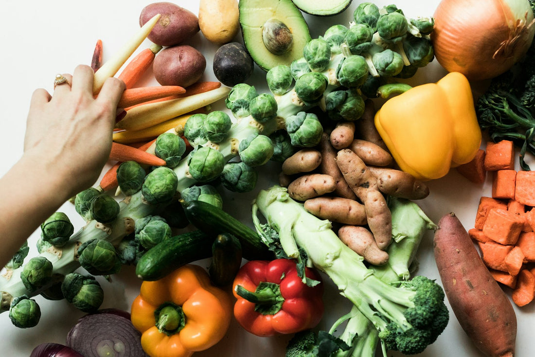 Fresh whole vegetables on a white surface, including peppers, broccoli, carrots, sweet potato and Brussels sprouts.