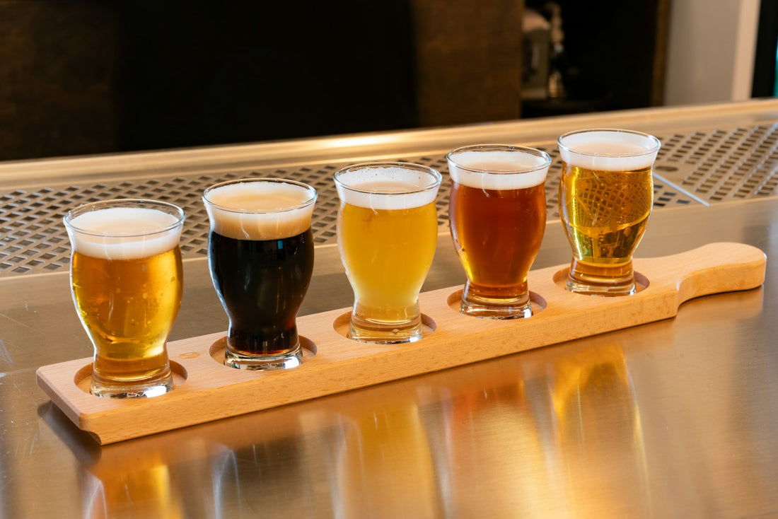 A row of five beer glasses of different colours on a wooden tasting paddle on a bright bar counter