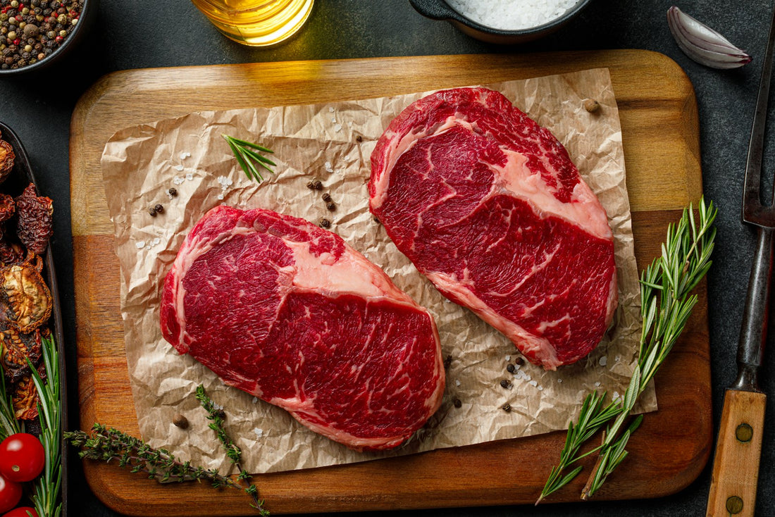 Two raw marbled beef steaks on a wooden board with rosemary and peppercorns