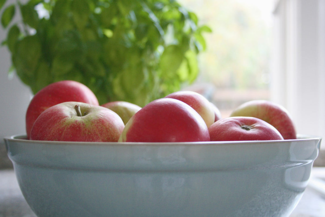 A white bowl of red and green apples on a table by a window