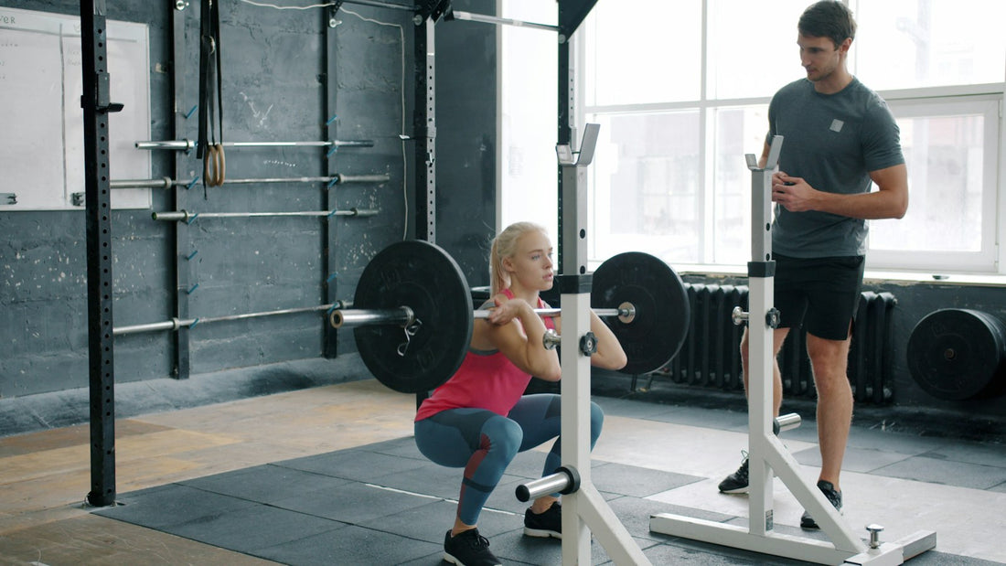 A person performing a barbell back squat in a gym while a coach watches.