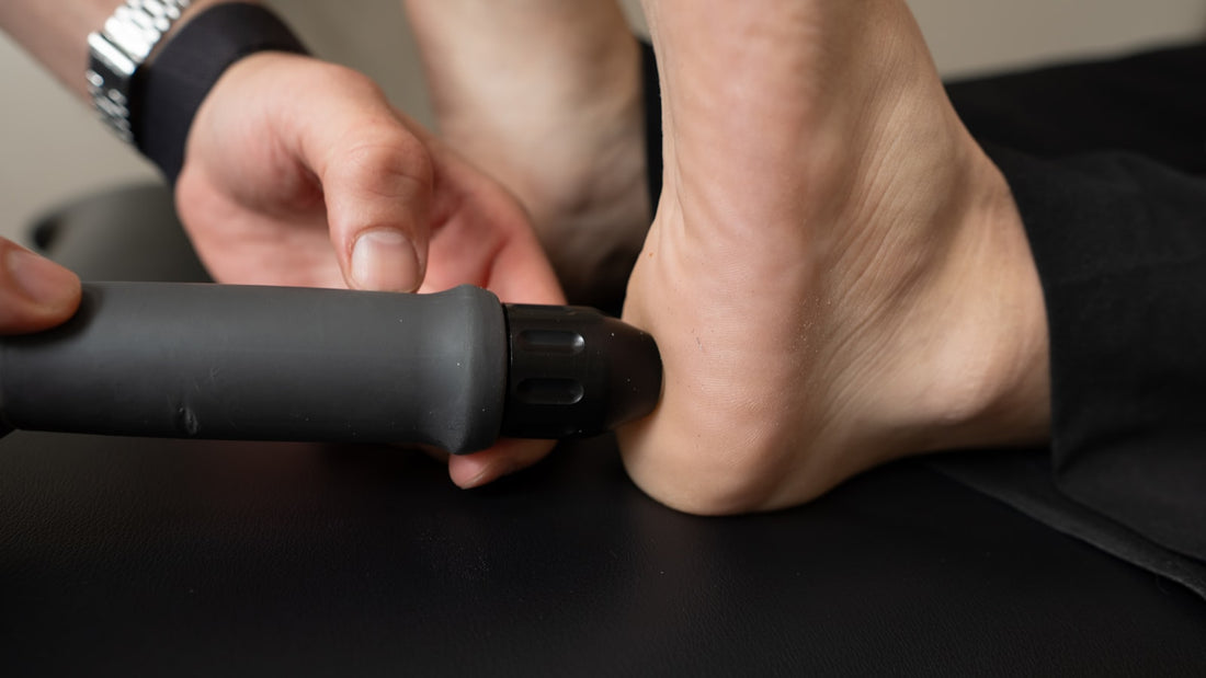 A handheld percussion massage device being applied to a person's lower leg above the ankle